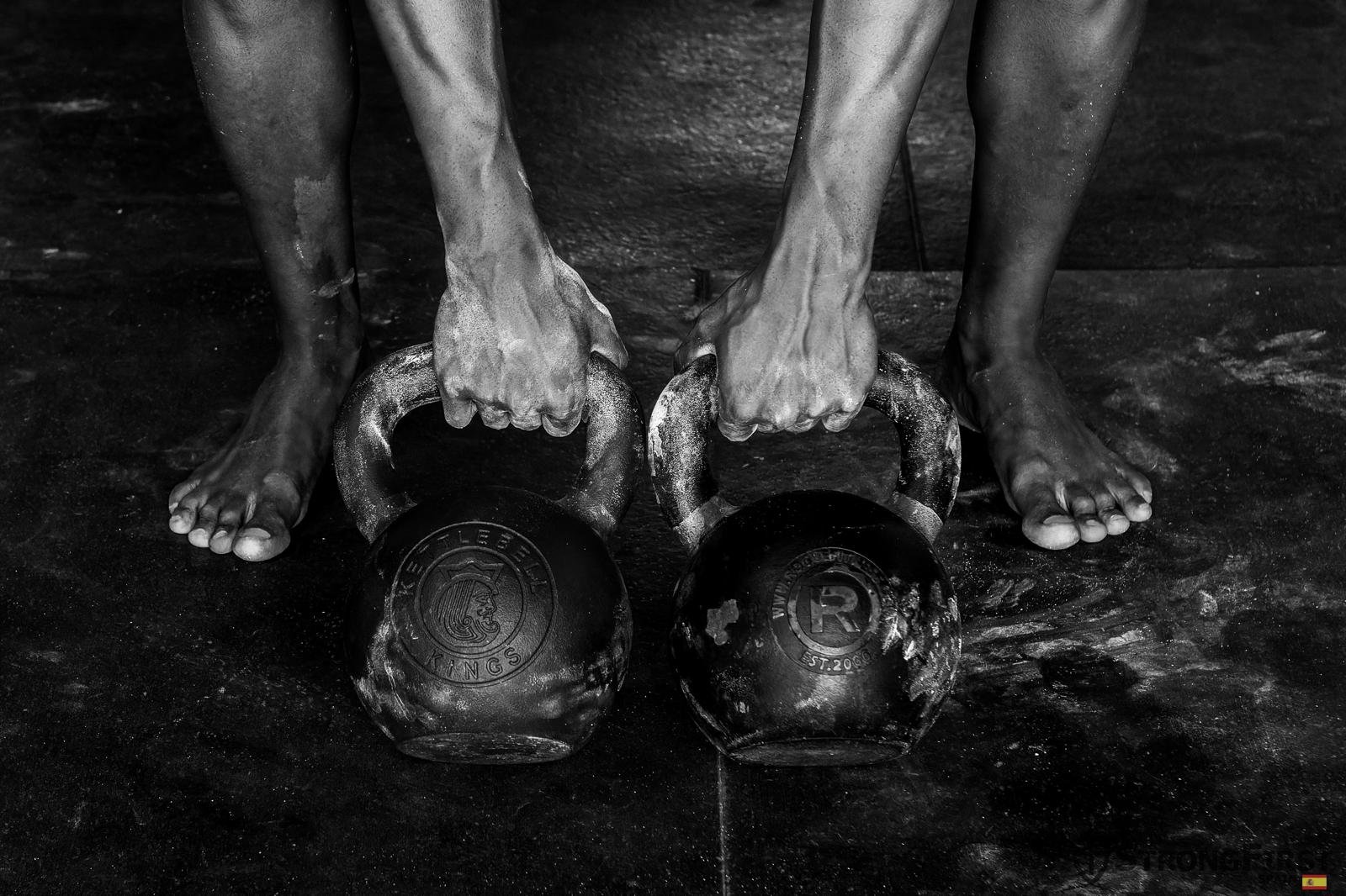 Mãos calejadas seguras nas pegas de dois kettlebells, pés descalços apoiados no chão